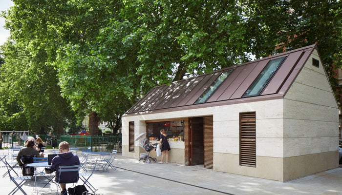 External shot of Brook Green Pavilion with leafy green trees behind and people sitting on outdoor furniture in front 