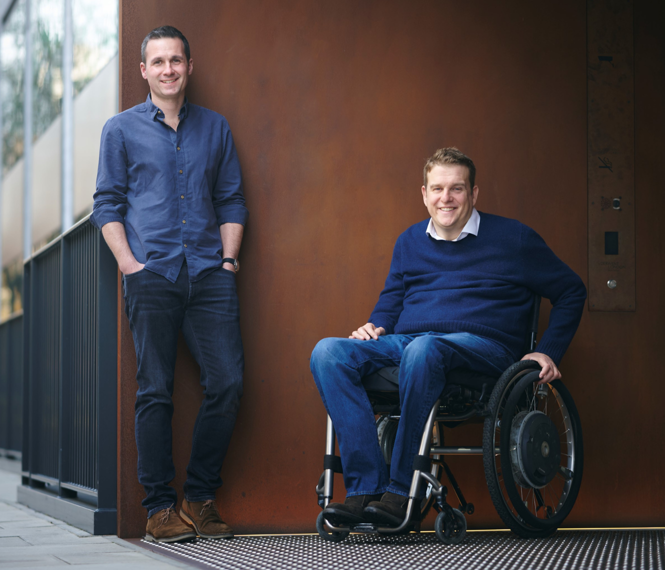 Ed Warner and James Taylor next to each other, outside a building. Ed standing, James sitting in his wheelchair